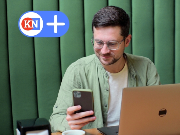 Student sitzt lächelnd vor einem Laptop und hat ein Handy in der Hand. Er konsumiert das Angebot von KN+.